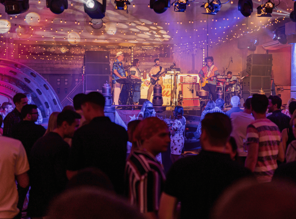 Volle Tanzfläche mit feiernden Gästen und einer Live-Band unter buntem Partylicht und Lichterketten.